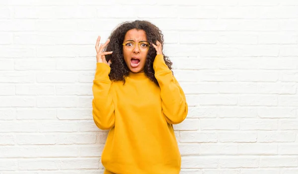 young black woman screaming with hands up in the air, feeling furious, frustrated, stressed and upset against brick wall