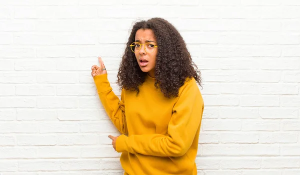 young black woman feeling shocked and surprised, pointing to copy space on the side with amazed, open-mouthed look against brick wall