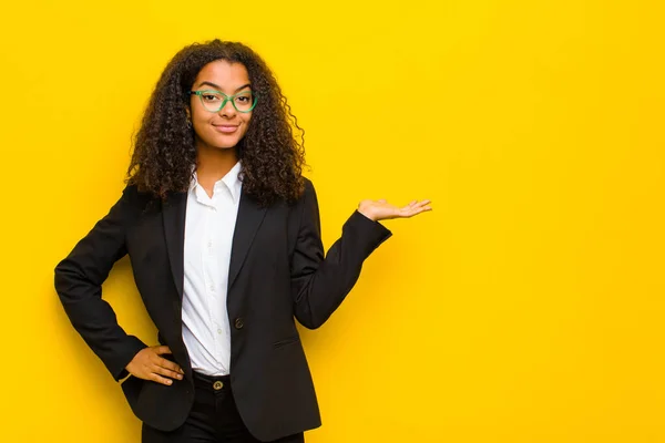 black business woman smiling, feeling confident, successful and happy, showing concept or idea on copy space on the side against orange wall