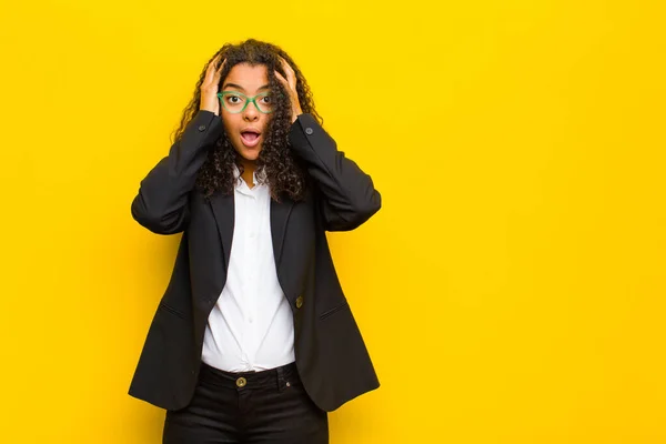 black business woman looking excited and surprised, open-mouthed with both hands on head, feeling like a lucky winner against orange wall