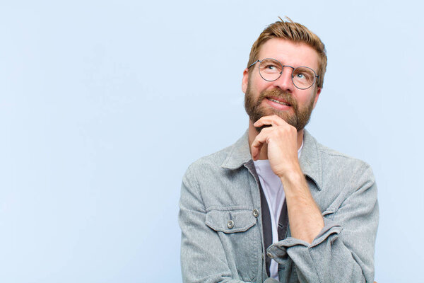 young blonde adult man feeling thoughtful, wondering or imagining ideas, daydreaming and looking up to copy space