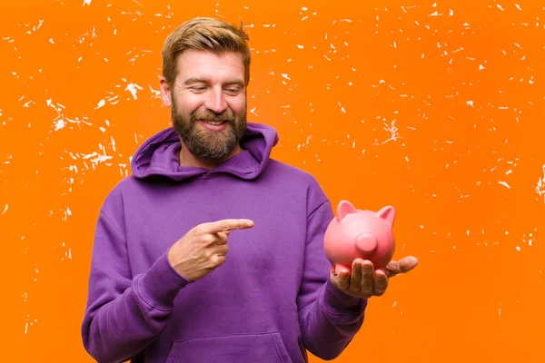 ダメージを受けたオレンジの壁に紫色のパーカーを着た貯金箱を持つ若いブロンドの男 — ストック写真