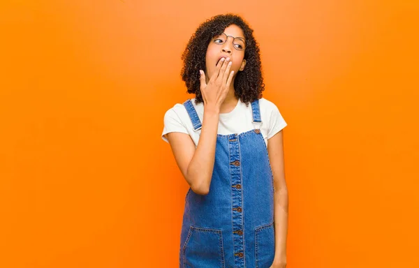 young pretty black woman yawning lazily early in the morning, waking and looking sleepy, tired and bored against orange wall