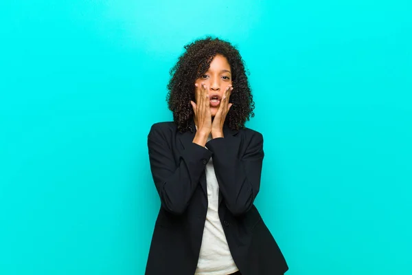 young black woman feeling shocked and scared, looking terrified with open mouth and hands on cheeks against blue wall