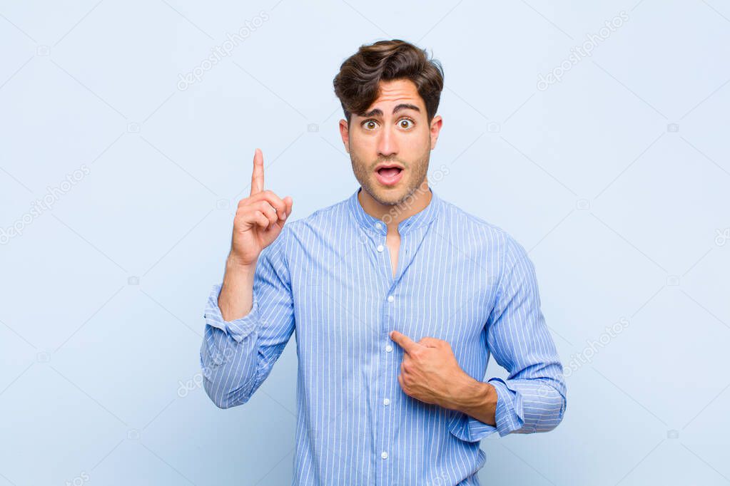 young handsome man feeling proud and surprised, pointing to self confidently, feeling like successful number one against blue background