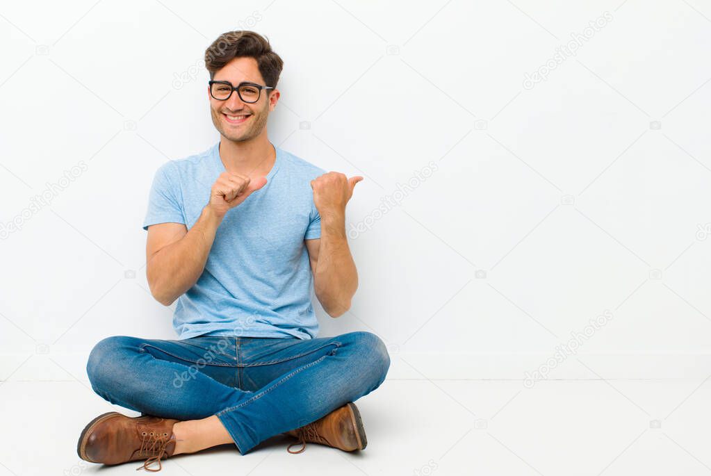 young handsome man smiling cheerfully and casually pointing to copy space on the side, feeling happy and satisfied sitting on the floor