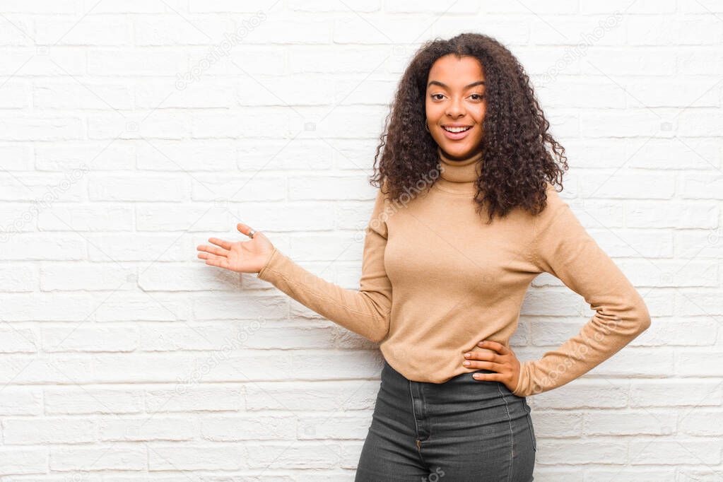 young black woman feeling happy and cheerful, smiling and welcoming you, inviting you in with a friendly gesture against brick wall