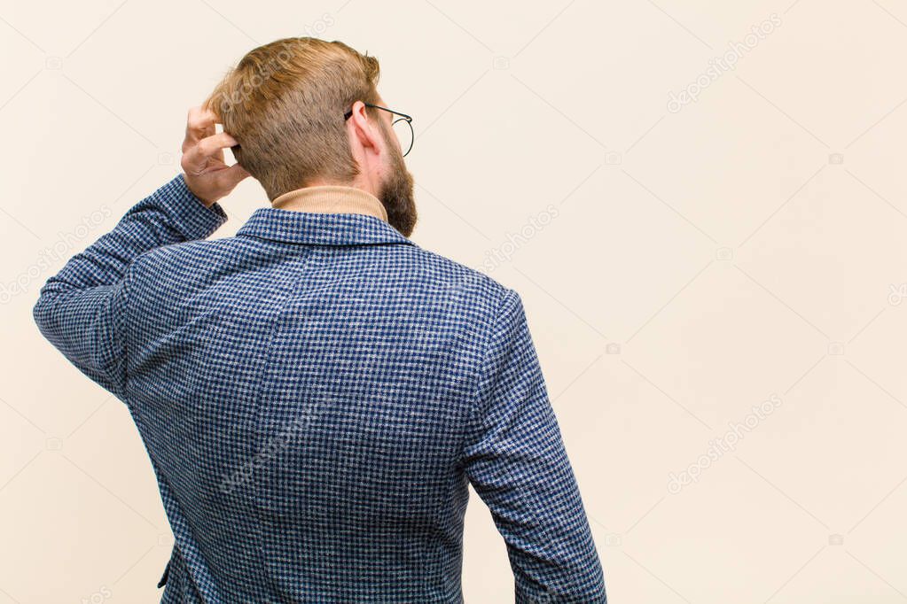 young blonde businessman feeling clueless and confused, thinking a solution, with hand on hip and other on head, rear view