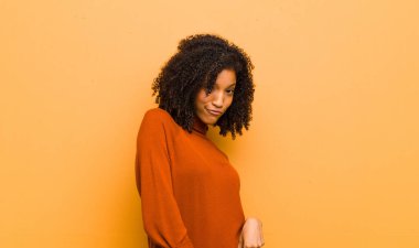 Genç, güzel siyah kadın gururlu, kendinden emin, havalı, küstah ve küstah görünüyor, gülümsüyor, turuncu duvara karşı başarılı hissediyor.