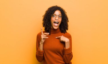 Genç ve güzel siyahi kadın gururlu ve şaşırmış hissediyor. Kendine güvenerek turuncu duvara karşı başarılı olduğunu hissediyor.