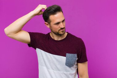 Genç yakışıklı adam şaşkın ve şaşkın hissediyor, başını kaşıyor ve mor duvara bakıyor.