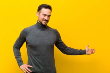 Genç yakışıklı adam kendini mutlu ve neşeli hissediyor, gülümsüyor ve seni selamlıyor, turuncu duvara karşı dostça bir jestle içeri davet ediyor.