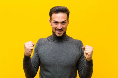 Genç yakışıklı adam şok olmuş, heyecanlı ve mutlu, gülüyor ve başarıyı kutluyor, vay canına! turuncu duvara yaslan