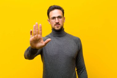 Genç yakışıklı adam ciddi, sert, hoşnutsuz ve kızgın görünüyor. Avuç içini açıp turuncu duvara karşı jest yapıyor.