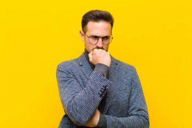 Genç yakışıklı adam ciddi, düşünceli ve endişeli, elleri turuncu duvara yaslanmış bir şekilde yan bakıyor.