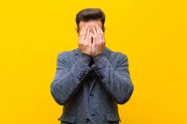 Genç yakışıklı adam üzgün, hayal kırıklığına uğramış, gergin ve depresif, yüzünü iki eliyle kapatmış, turuncu duvara karşı ağlıyor.