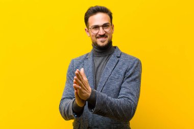 Genç yakışıklı adam mutlu ve başarılı hissediyor, gülümsüyor ve el çırpıyor, turuncu duvara karşı alkış tutarak tebrik ediyor.