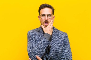 Genç yakışıklı adam şok ve inançsızlık içinde, elleri yanak ve kolları çaprazlanmış, sersemlemiş ve turuncu duvara karşı şaşkın hissediyordu.