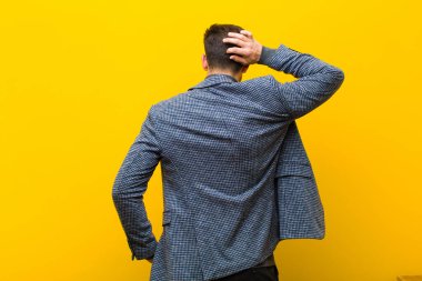 Genç yakışıklı adam düşünüyor ya da şüphe ediyor, kafasını kaşıyor, şaşkın ve kafası karışık hissediyor, turuncu duvarın arkasında ya da arkasında.