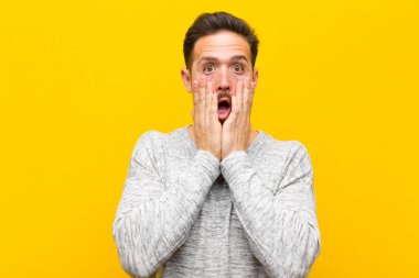 Genç yakışıklı adam şok olmuş ve korkmuş hissediyor. Ağzı açık ve elleri turuncu duvarda.