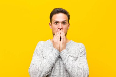 Genç yakışıklı adam endişeli, üzgün ve korkmuş hissediyor, elleriyle ağzını kapatıyor, endişeli görünüyor ve turuncu duvara karşı berbat durumda.