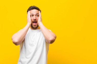 Genç yakışıklı adam mutlu, heyecanlı ve şaşırmış, yüzünü turuncu duvara dayamış, yan tarafa bakıyor.