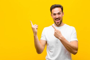Genç yakışıklı adam mutlu ve şaşırmış hissediyor, şok olmuş bir ifadeyle gülümsüyor ve turuncu duvara işaret ediyor.
