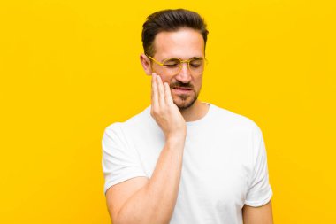 Genç yakışıklı adam yanağını tutuyor ve ağrılı diş ağrısı çekiyor. Hasta, sefil ve mutsuz hissediyor. Turuncu duvarda dişçi arıyor.