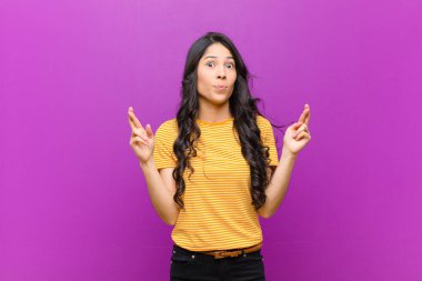 Genç, güzel, Latin bir kadın endişeyle dua ediyor. Mor duvara karşı endişeli bir bakışla iyi şanslar diliyor.