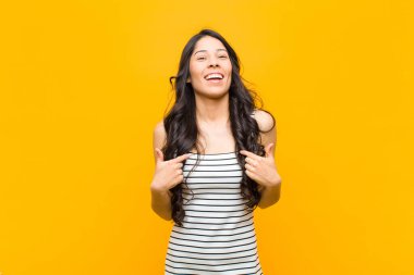 Genç, güzel, Latin bir kadın mutlu, şaşırmış ve gururlu hissediyor. Heyecanlı ve şaşkın bir şekilde turuncu duvara bakıyor.