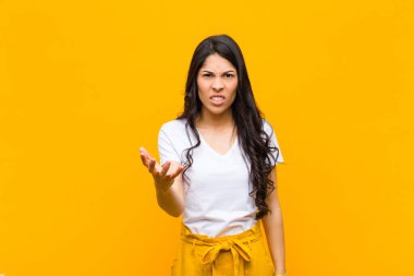 Genç, güzel, Latin bir kadın kızgın, sinirli ve sinirli görünüyor. Portakal duvarına karşı senin neyin var? 