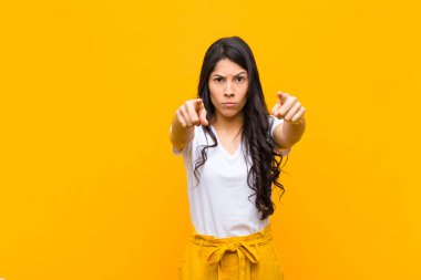 Genç, güzel, Latin bir kadın kameraya hem parmaklarıyla hem de kızgın bir ifadeyle bakıyor. Turuncu duvara karşı görevini yapmanı söylüyor.