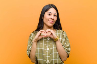 Genç ve güzel İspanyol kadın gülümsüyor ve mutlu, sevimli, romantik ve aşık, iki elini de kahverengi duvara dayayarak kalbi şekillendiriyor