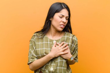 Genç, güzel İspanyol kadın üzgün, yaralı ve kalbi kırık görünüyor, iki elini de kalbe yakın tutuyor, ağlıyor ve kahverengi duvara karşı depresif hissediyor.