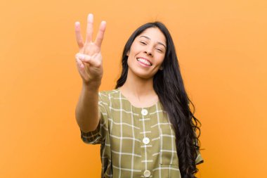 Genç, güzel İspanyol kadın gülümsüyor ve arkadaş canlısı görünüyor. Üç ya da üç numarayı gösteriyor. Elleri önde, kahverengi duvara doğru sayıyor.