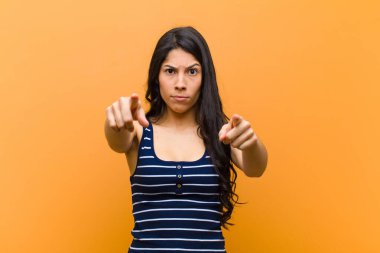 Genç, güzel, İspanyol bir kadın kameraya doğru bakıyor. Hem parmakları hem de kızgın bir ifadeyle. Kahverengi duvara karşı görevini yapmanı söylüyor.