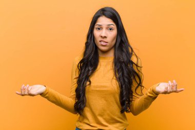 Genç, güzel, İspanyol bir kadın kendini cahil ve kafası karışık hissediyor. Hangi seçeneği seçeceğinden emin değil. Kahverengi duvara karşı merak içinde.