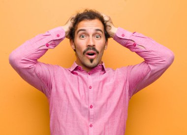 Genç yakışıklı adam heyecanlı ve şaşırmış görünüyor, ağzı açık, iki eli başının üstünde, turuncu duvara karşı kendini şanslı hissediyor.