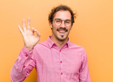 Genç yakışıklı adam kendini başarılı ve tatmin olmuş hissediyor, ağzı açık gülümsüyor, elini turuncu duvara dayayıp işaret veriyor.