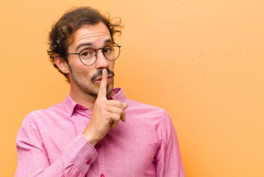 Genç yakışıklı adam sessizlik ve sessizlik istiyor, parmağıyla ağzının önünde işaret ediyor, ya da turuncu duvara karşı bir sır saklıyor.