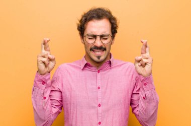 Genç yakışıklı adam gülümsüyor ve endişeyle iki parmağını çapraz yapıyor. Endişeli ve turuncu duvara karşı iyi şanslar diliyor ya da umut ediyor.