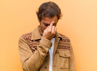 Genç yakışıklı adam stresli, mutsuz ve hayal kırıklığına uğramış, alnına dokunuyor ve turuncu duvara karşı şiddetli baş ağrısı çekiyor.