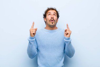Genç ve yakışıklı bir adam korkuyla ve açık ağızla yukarıyı işaret ediyor. Şaşırmış ve şaşırmış görünüyor. Düz duvara yaslanmış.