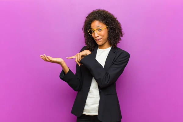 young black business woman smiling cheerfully and pointing to copy space on palm on the side, showing or advertising an object