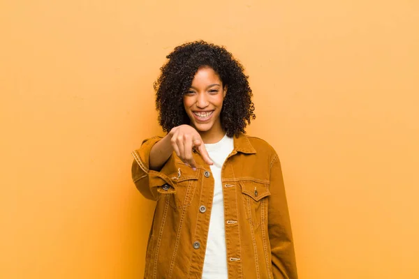 young pretty black woman pointing at camera with a satisfied, confident, friendly smile, choosing you against orange wall