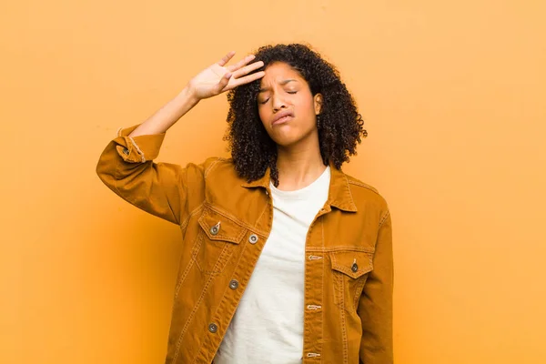 young pretty black woman looking stressed, tired and frustrated, drying sweat off forehead, feeling hopeless and exhausted against orange wall