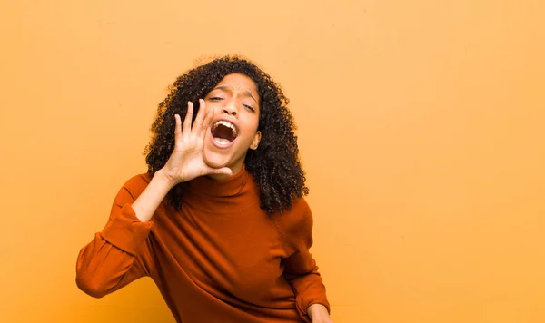 young pretty black woman profile view, looking happy and excited, shouting and calling to copy space on the side against orange wall