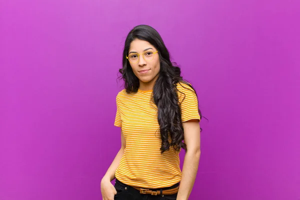 young pretty latin woman smiling cheerfully and casually with a positive, happy, confident and relaxed expression against purple wall