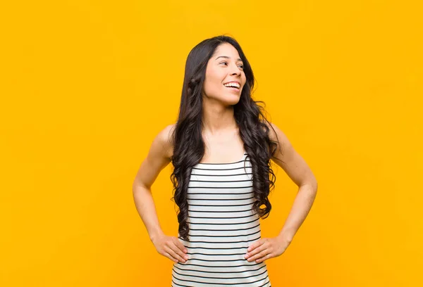 young pretty latin woman looking happy, cheerful and confident, smiling proudly and looking to side with both hands on hips against orange wall