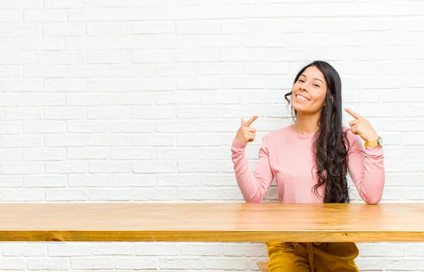 young  pretty latin woman smiling confidently pointing to own broad smile, positive, relaxed, satisfied attitude sitting in front of a table